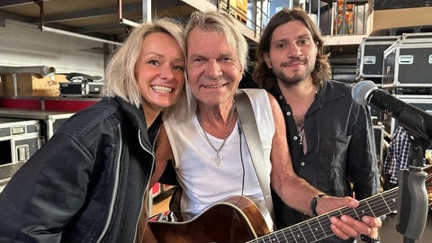 Matthias Reim steht mit Gitarre um den Hals zwischen Ehefrau Christin Stark und Sohn Julian Reim. Sie lachen in die Kamera.